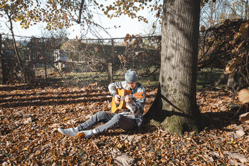 young man in a plaid shirt and beanie sits under a large tree, playing an acoustic guitar in a sunny autumn setting. The ground is covered with dry leaves, enhancing the cozy and peaceful atmosphere