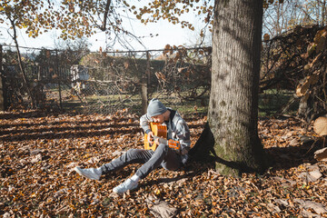 young man in a plaid shirt and beanie sits under a large tree, playing an acoustic guitar in a sunny autumn setting. The ground is covered with dry leaves, enhancing the cozy and peaceful atmosphere