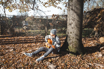 young man in a plaid shirt and beanie sits under a large tree, playing an acoustic guitar in a sunny autumn setting. The ground is covered with dry leaves, enhancing the cozy and peaceful atmosphere