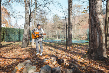Guitarist in a plaid shirt stands confidently on a pile of stones in an autumn park, strumming his...