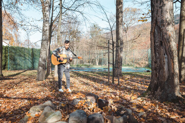 Guitarist in a plaid shirt stands confidently on a pile of stones in an autumn park, strumming his...