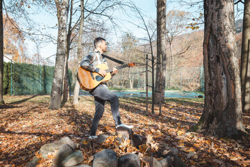 Guitarist in a plaid shirt stands confidently on a pile of stones in an autumn park, strumming his...