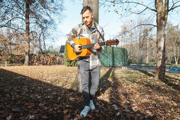 Young man leaning against a tree, playing an acoustic guitar in a sunlit autumn park, surrounded by...