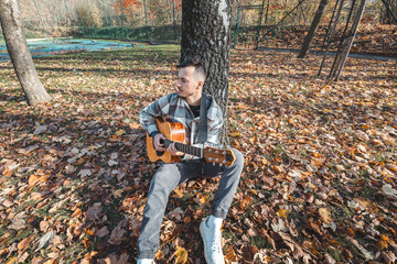 Man in a plaid shirt tunes his acoustic guitar while sitting on autumn leaves in a serene park. Vibrant fall colors and natural ambiance enhance the creative mood