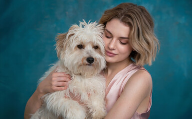 A young woman with wavy, shoulder-length hair, gently embracing a fluffy white dog. They are positioned against a textured blue backdrop. 