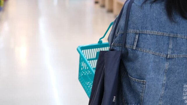 A person is holding a shopping cart with a blue basket and a blue bag. The shopping cart is in a store, and the person is walking down the aisle