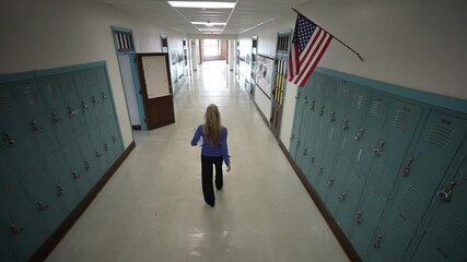 A mature woman wearing casual attire walks down a school hallway lined with lockers. An American flag hangs above, enhancing the educational atmosphere.