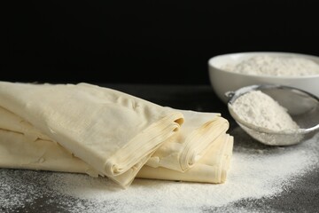 Raw puff pastry dough and flour on dark table against black background, closeup