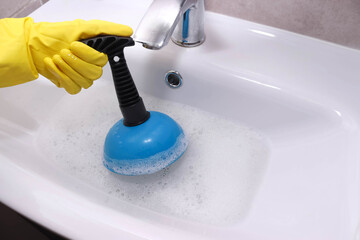 Hand in yellow gloves cleaning sink with plunger. Overflowing bathroom sink. Clogged drain.