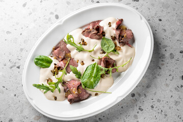 A elegant dish featuring sliced roast beef topped with a creamy sauce, garnished with fresh greens and crunchy toppings, presented on a white plate against a textured background.