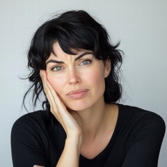 A woman with black, wavy hair gazes pensively, resting her chin on her hand. She displays a contemplative expression, surrounded by a soft, neutral background