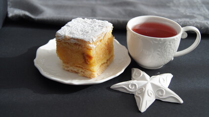 Sponge cake with cream, English soup cake sprinkled generously with powdered sugar, a piece of cake with vanilla cream and impregnation, dessert with a cup of fruit cherry tea, on a black background