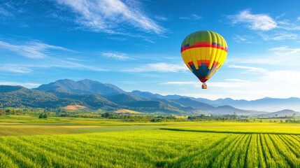 Obraz premium A colorful hot air balloon flying over a green field with mountains in the background. Represents travel, adventure, freedom, and scenic landscapes.