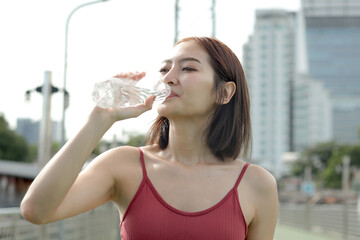 Short haired sporty girl in sportswear holds water bottle while takes a break after jogging work out to exercise outdoor on city street. Happy smiling Asian woman drinking water to refresh herself.