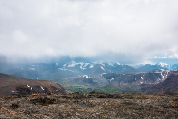 Dramatic top view from sunlit stony pass to vivid green alpine valley in sunlight against large mountain range with snow-capped peaks in rainy low clouds. Misty snowy mountain tops in rain cloudy sky.