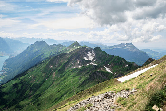 Stunning panoramic view of the Swiss Alps near Brienz, featuring lush green slopes, rocky trails, and snowy peaks under a partly cloudy sky.