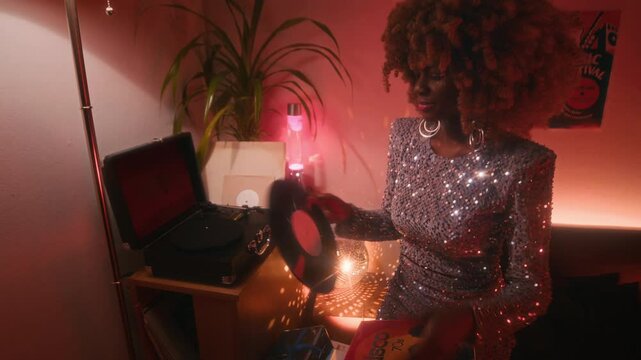 Medium low key shot of fashionable young African American disco star with puffy hair, wearing shiny silver dress, playing 70s vinyl record in record player in neon lit room