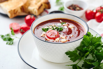 Tomato puree soup garnished with parsley, feta cheese and spices with toasted bread slices, tomatoes and herbs on white table