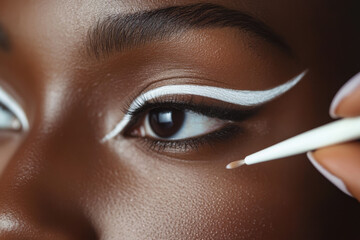 A makeup artist applies precise white eyeliner to the eye of a model with dark skin. The artistic design adds flair and highlights beauty during an artistic session.