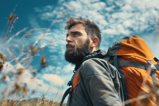 Man with a beard and a backpack is standing in a field. He looks up at the sky and seems to be enjoying the outdoors