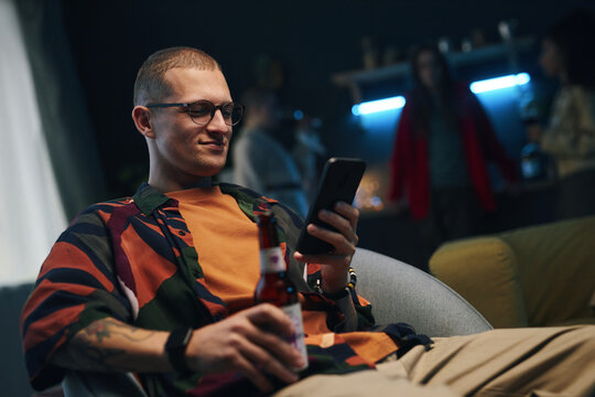 Young man drinking beer and surfing the net in smartphone while sitting on sofa at home party