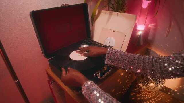 Low key closeup of hands of unrecognizable lady in shiny silver dress taking phonograph record and placing it on retro vinyl record player in neon lit room with disco ball
