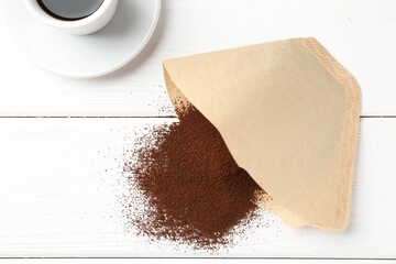 Paper filter, ground coffee and cup of drink on white wooden table, flat lay
