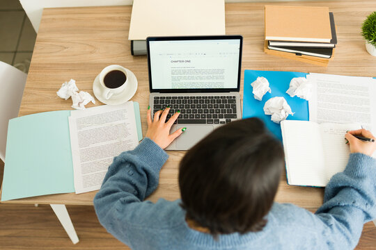 Top view of  a young woman writing a book at home, using laptop and notebook, with crumpled papers and coffee cup on desk