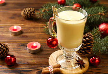 Tasty Christmas cocktail (eggnog) in glass, festive decor and burning candles on wooden table, closeup