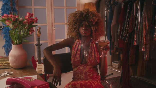 Medium portrait of 70s young Black female celebrity with puffy hair, sparkling jewelry on neck, in open hippie dress, sitting at dressing table with glass of martini and looking calmly at camera