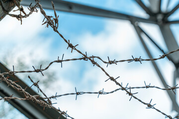 Barbed wire stretches across a blue sky with clouds near an industrial structure in a rural area