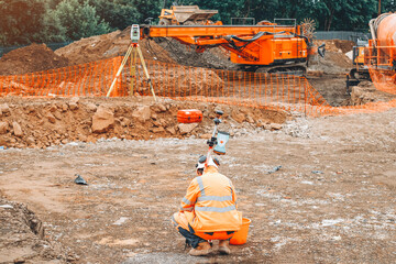 Construction site engineer surveying land at a development site surrounded by machinery and...
