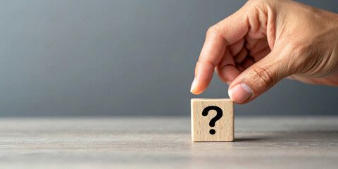 A hand gently holds a wooden block with a question mark printed on it against a plain background. The block rests on a smooth, light-colored surface. AI generated.