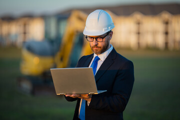 Civil engineer worker at a construction site. Mature engineer worker. Man in suit and hardhat helmet at construction site. Middle aged head civil engineer worker standing outside near excavator.