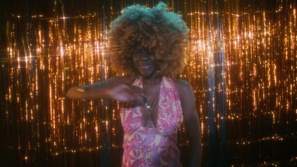 Medium low key portrait of young African American lady with puffy hair wearing colorful hippie open dress, dancing actively and charismatically against glittery tinsel background in club, 70s