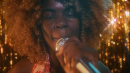 Close-up low key portrait of young African American lady with puffy hair singing sensually into microphone on glittery golden background in night club and looking at camera