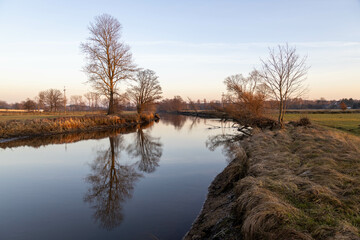 The Świder River Reserve on a winter afternoon in Eastern Europe without snow cover, trees without leaves reflected in the water, a calm river