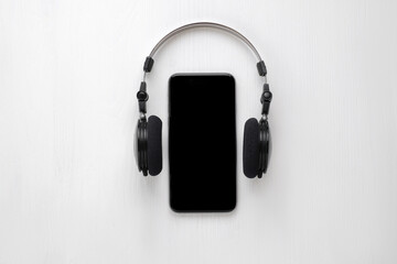 A smartphone with a black screen placed on a white surface, framed by black over-ear headphones.
