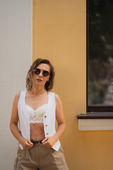 Stylish woman in sunglasses and a white sleeveless vest stands against a yellow wall. She is wearing white lace lingerie and beige shorts, adding a chic touch to her street-style look