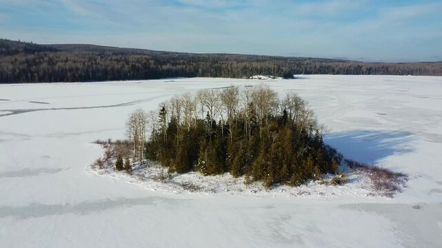 Drone orbit around a remote wooded island surrounded by a vast frozen lake in a cold winter landscape of Lake Matapedia, Matapedia Valley, Quebec, Canada.