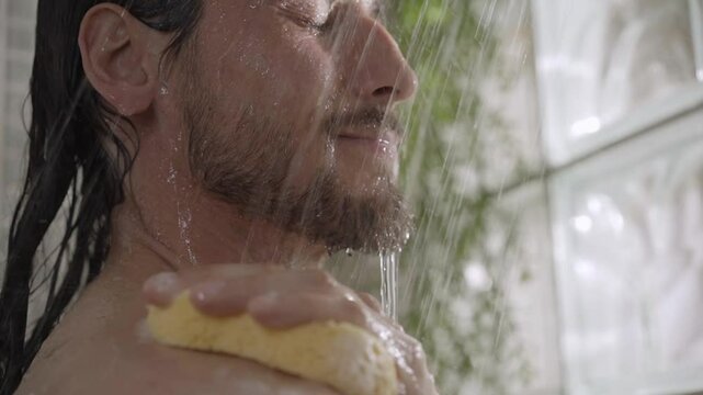 Young long hair man rinsing soap from skin sponge in bathroom. Close-up of adult Caucasian bearded male performing daily personal care and hygiene activity alone in shower at home