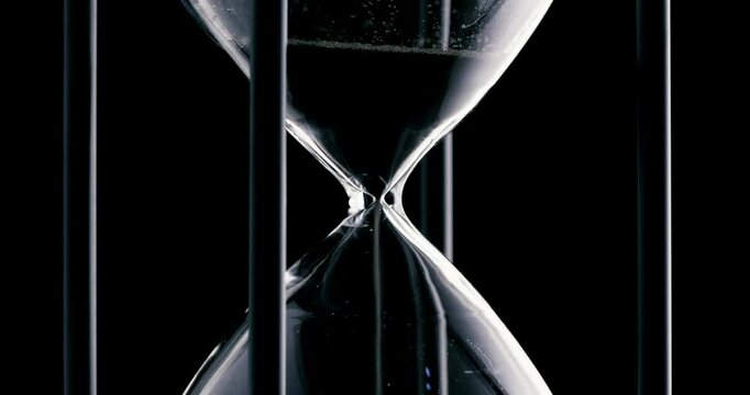 Rotation of sand timer clock with sand flowing from through hourglass on black background.
