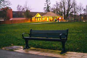 A bench in the foreground with an inviting café glows warmly in the dim light of a winter evening,...