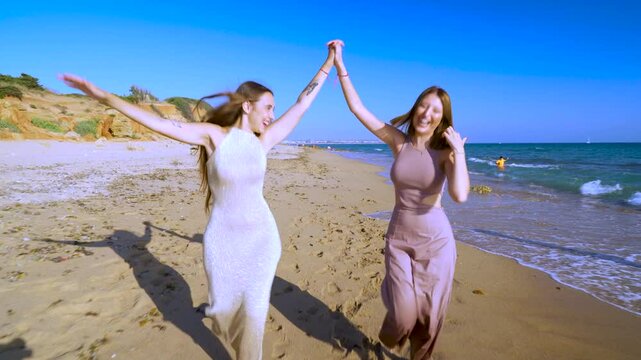 Dos chicas j&oacute;venes corriendo y sonriendo muy divertidas, por la orilla de la playa sobre la arena junto al mar, un magn&iacute;fico d&iacute;a soleado con cielo azul.