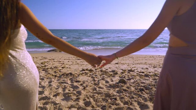 Dos mujeres cogidas de la mano andan sobre la arena de la playa hacia el mar un d&iacute;a soleado con cielo azul.