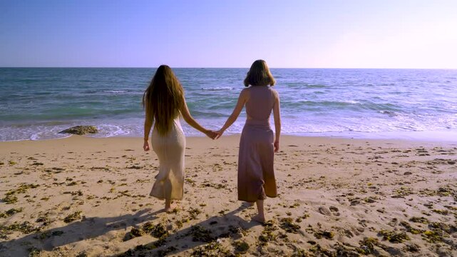 Dos mujeres cogidas de la mano andan sobre la arena de la playa hacia el mar un d&iacute;a soleado con cielo azul.