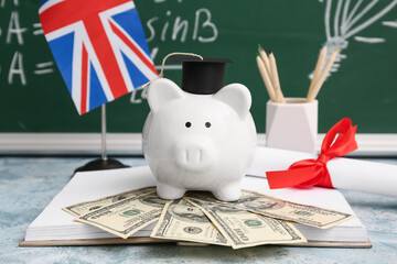 Piggy bank with money, diploma, book and UK flag on table near blackboard, closeup. Student loan...