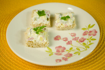 Four square pieces of bread topped with creamy egg salad and parsley, served on a plate with delicate pink floral patterns, placed on a vibrant yellow mat.