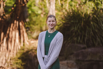 A smiling woman in a green dress with a gray cardigan stands outdoors, framed by lush green plants and soft sunlight, creating a serene, natural atmosphere © Keely Anne