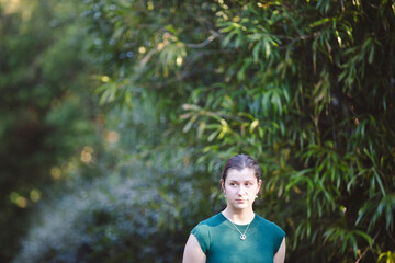 A woman in a sleeveless green dress stands outdoors, looking at the camera, with dense greenery and...